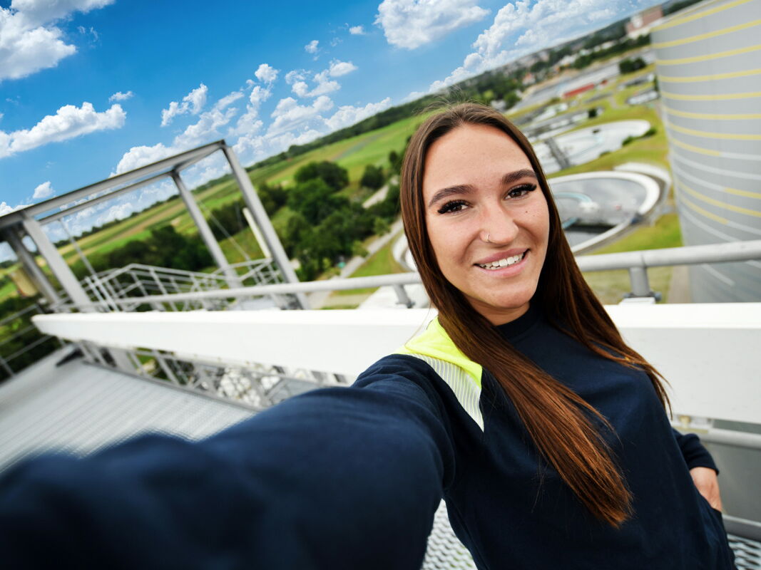 Junge Frau macht ein Selfie von sich auf einer Anlagenbrücke mit Klärbecken im Hintergrund