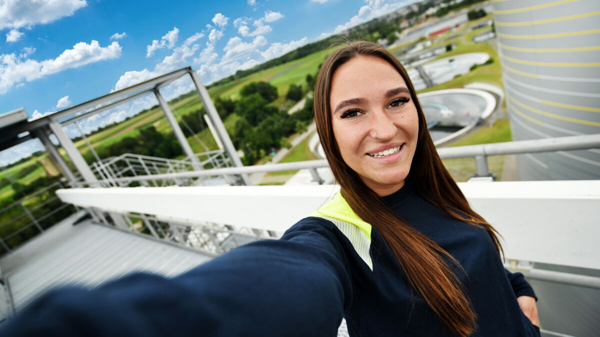 Junge Frau macht ein Selfie von sich auf einer Anlagenbrücke mit Klärbecken im Hintergrund