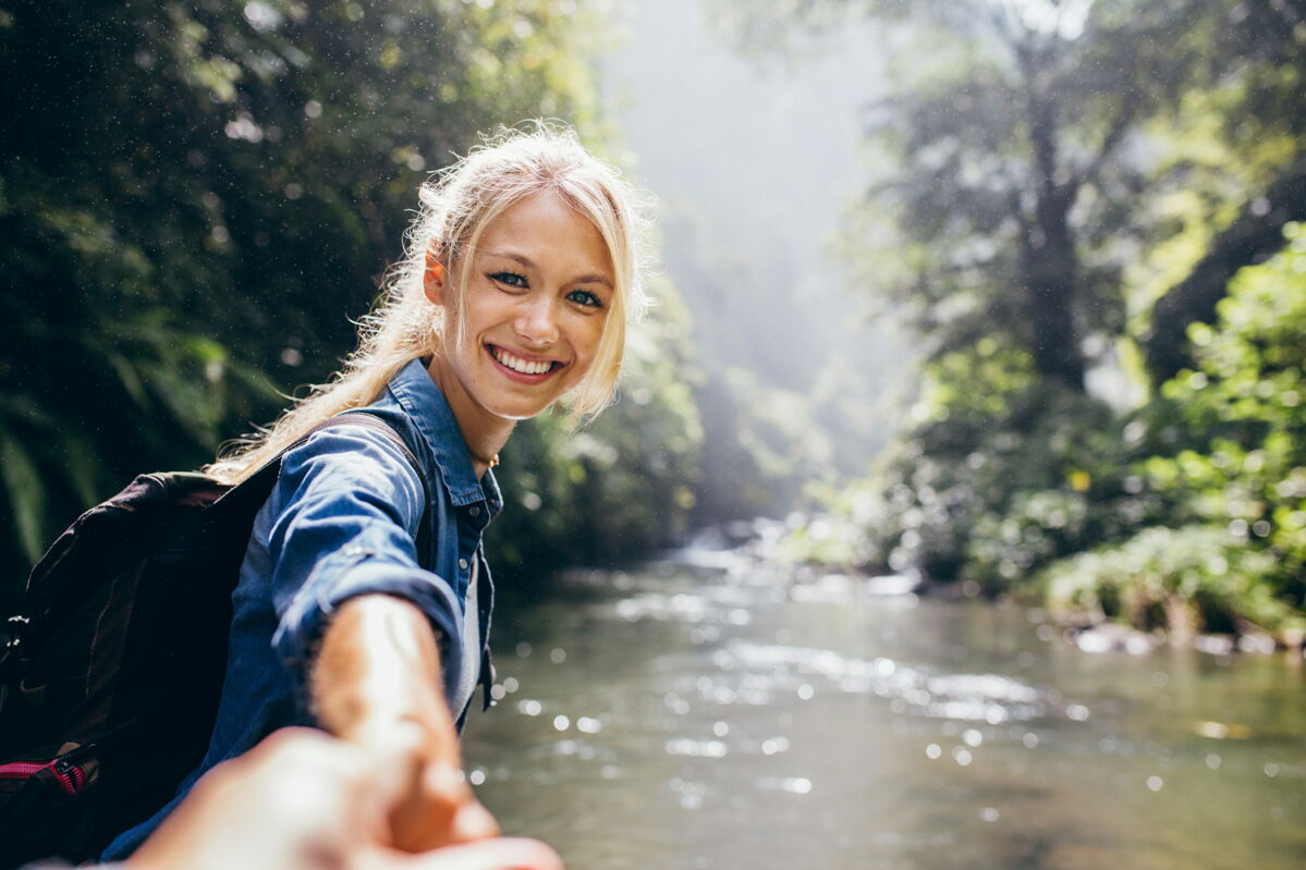 Portrait einer glücklichen jungen Frau, die die Hand ihres Freundes hält beim Wandern an einem Fluss